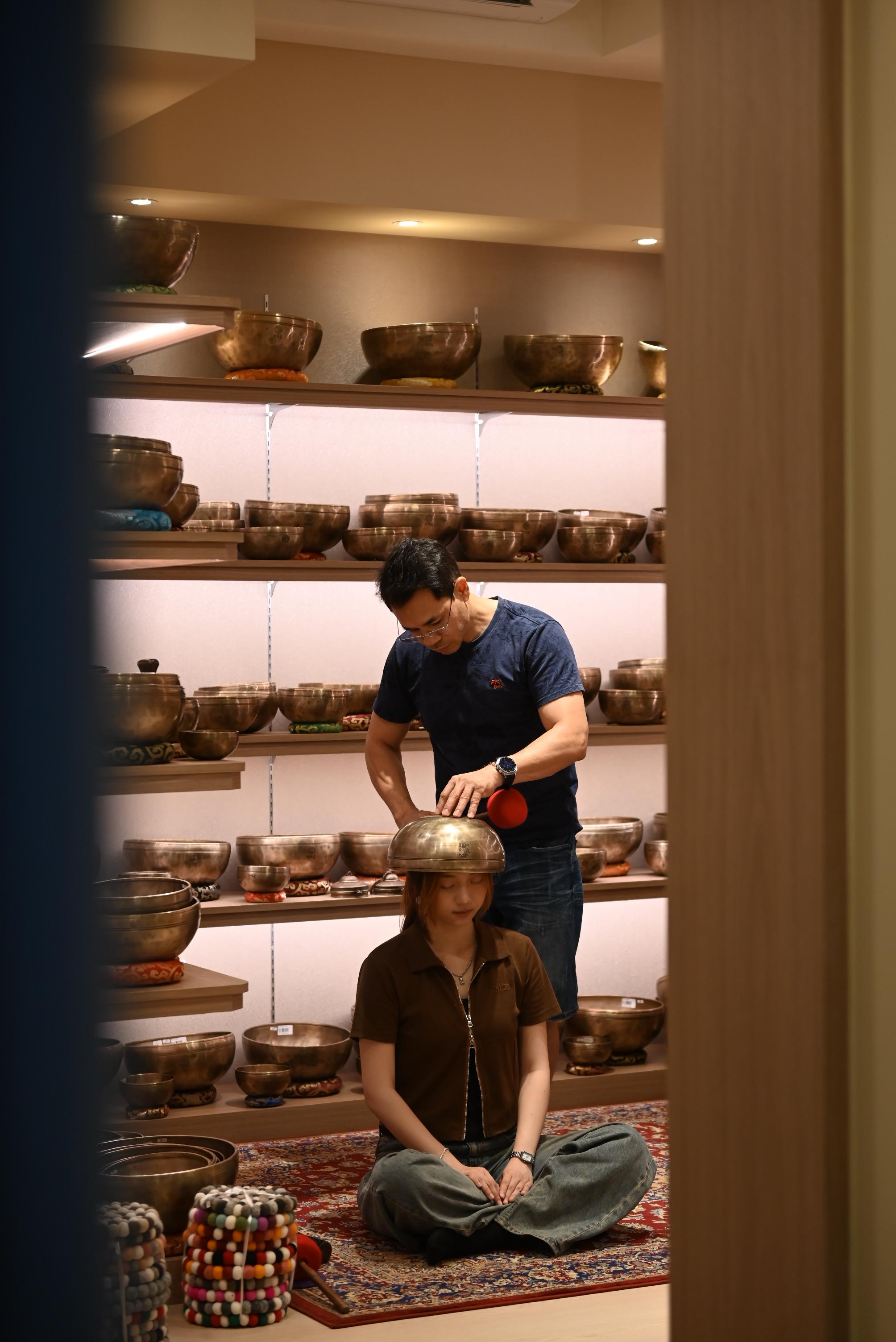 A man lowers a singing bowl onto the chest of a woman lying on a Persian rug, warm-lit shelves of bronze bowls behind them