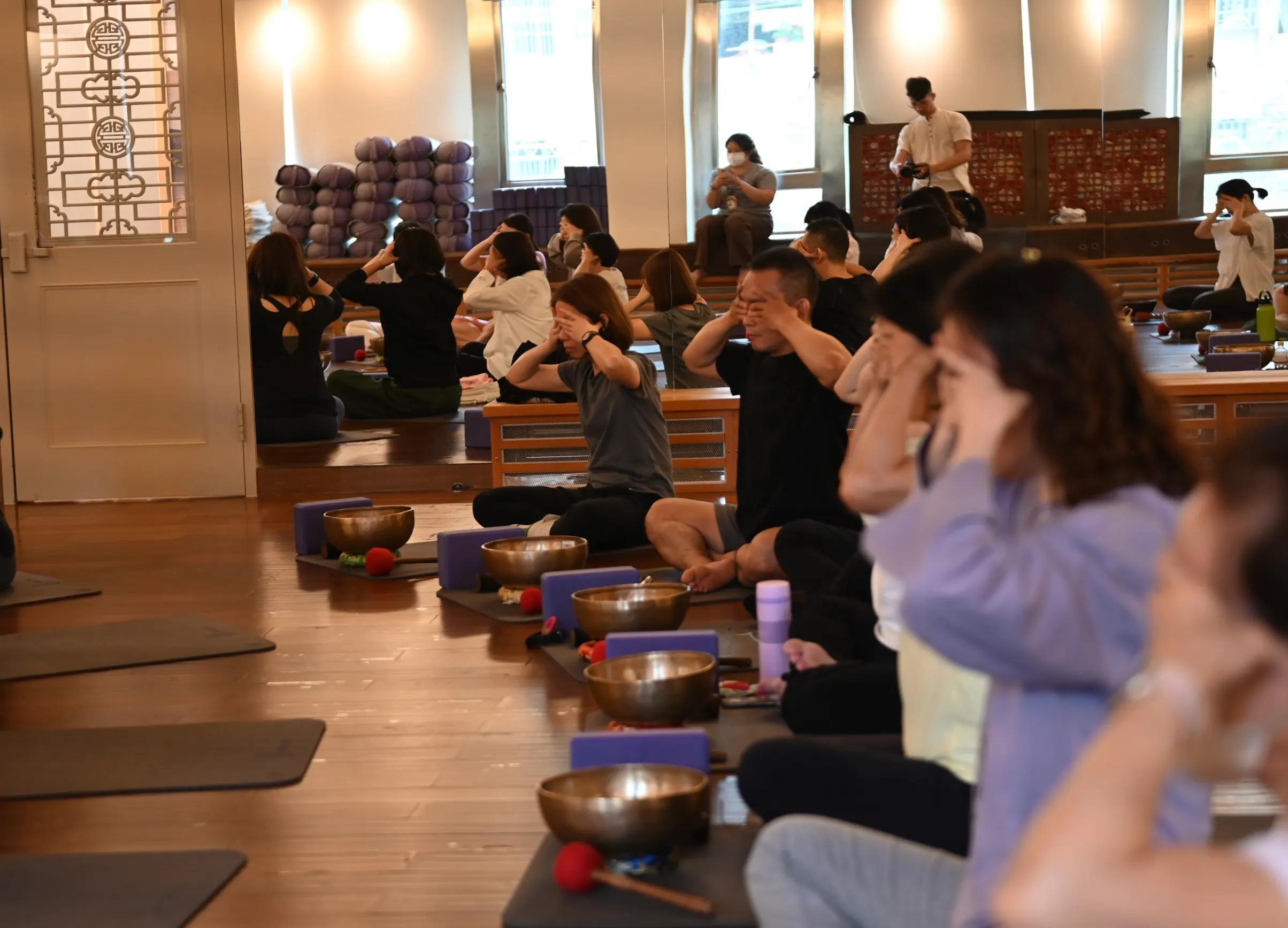 About fifteen people sit cross-legged on mats in a mirrored wood-floored hall with their hands over their eyes, bowls and purple blocks in front of them