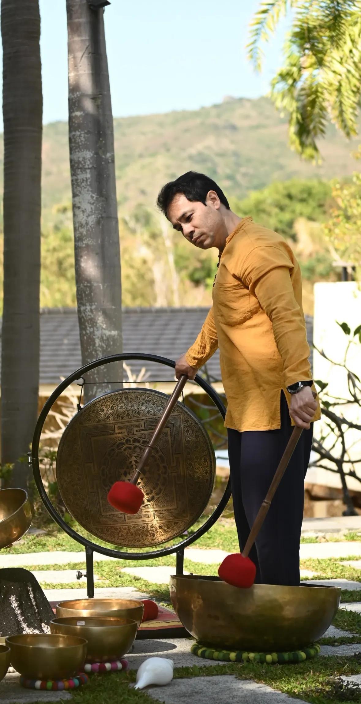 A man in a mustard kurta strikes a large engraved gong on a stand in a sunlit garden, a bowl and mallets on the stone paving beside him