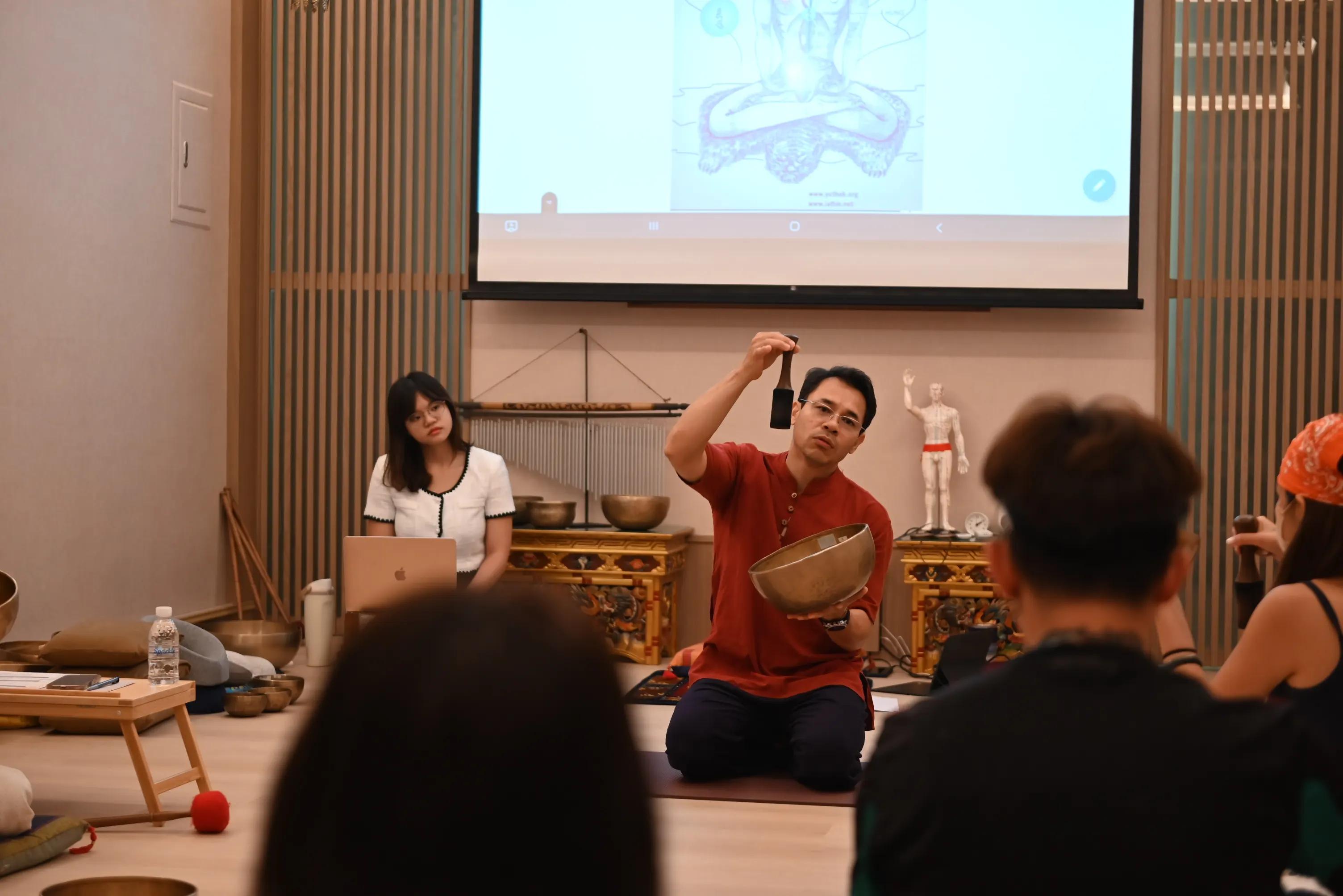 An instructor in a rust kurta kneels holding a bowl and raised mallet, teaching past the out-of-focus heads of students, a projected chakra anatomy slide behind